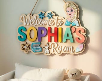 Può includere: Cartello in legno con la scritta "Welcome to SOPHIA'S Room" in lettere colorate, con un'illustrazione di Elsa di Frozen. Il cartello include disegni di fiocchi di neve e forziere del tesoro ed è appeso sopra una culla con un orsacchiotto.