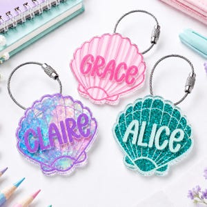 May include: Three seashell-shaped keychains with metal wire loops. One is pink with the name GRACE, one is purple with iridescent glitter and the name CLAIRE, and one is teal with glitter and the name ALICE. Each features raised lettering.