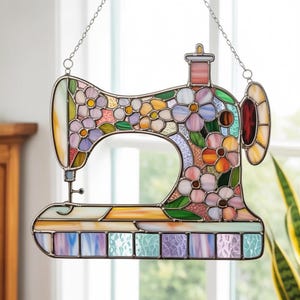 Può includere: Macchina da cucire in vetro colorato con design floreale. Il pezzo colorato presenta una varietà di fiori nelle tonalità del rosa, viola, arancione e giallo. La base ha una fila di pannelli quadrati dai colori pastello. Una catena è attaccata per appenderla.