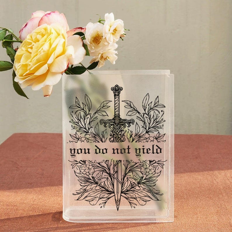 May include: Clear rectangular vase with a black sword and floral design, and the text "you do not yield". The vase is filled with yellow and white roses, set on a pink surface.