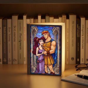 May include: A decorative book-shaped lamp featuring a stained-glass design with Hercules and Megara from the Disney movie. The lamp is illuminated, showcasing the colorful artwork. It is set on a wooden surface with books in the background.