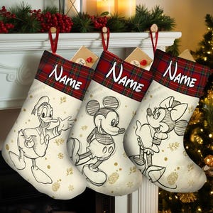 May include: Three Christmas stockings with Disney characters Donald Duck, Mickey Mouse, and Minnie Mouse. Each stocking has a red and green plaid cuff with the word "Name" in white. The stockings are cream-colored with gold snowflake and pinecone designs.