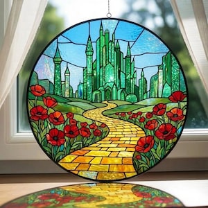 May include: Round stained glass panel depicting the Emerald City from The Wizard of Oz. The artwork features a yellow brick road, red poppies, and a green city under a blue sky. The panel is framed in black and hangs from a chain.