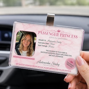 Könnte beinhalten: Eine rosa-weiße „Passenger Princess“-Offizielldokumentenkarte mit einem Foto einer Frau. Die Karte enthält den Text „Ausgestellt an Antonesha Staley“ und „Gültig bis in alle Ewigkeit“. Die Karte wird vor dem Armaturenbrett eines Autos gehalten.