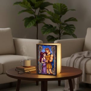 May include: A decorative book-shaped lamp with a stained-glass design featuring a cartoon couple. The lamp is illuminated, casting a warm glow. The image on the lamp depicts a man and woman in a colorful, classical style. The lamp is on a wooden table.