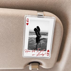 May include: Clear plastic car visor clip featuring a black and white photo of a couple on a beach. The design includes a Queen of Hearts playing card motif with the text "How did I get so Lucky?" in red script.