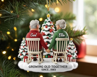 May include: A Christmas ornament featuring two elderly figures sitting in white rocking chairs, facing a decorated tree. The figure on the left wears a red jumper labelled Carl, and the right wears a green jumper labelled Ellie. Text reads: GROWING OLD TOGETHER SINCE...1983.