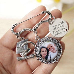 Op de afbeelding: Zilveren herdenkingsbroche met veiligheidsspeldsluiting. Het heeft een ronde hanger met een foto, een bedel met afstudeerhoed en een schijf met de tekst "I know you'd be with me today if heaven weren't so far away."