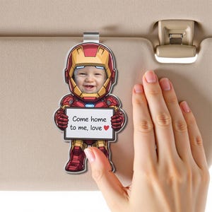 Puede incluir: Un clip personalizado para la visera del coche con la cara de un bebé dentro de un diseño de Iron Man. El clip es rojo, dorado y plateado, y sostiene un letrero que dice "Come home to me, love" con un símbolo de corazón.
