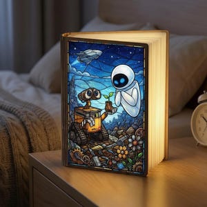 Puede incluir: Lámpara de madera con forma de libro con una colorida ilustración de Wall-E y Eva. La lámpara está abierta, con la obra de arte iluminada visible. La obra representa un robot sosteniendo una planta, con una nave espacial en el fondo. La lámpara está sobre una mesita de noche de madera.