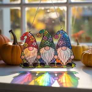 Puede incluir: Un adorno de ventana de vidrieras con tres gnomos con coloridos sombreros florales y largas barbas blancas. Los gnomos están sentados sobre una base verde, con una cadena para colgar. Frente a una ventana con calabazas.