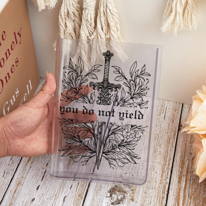 May include: Clear plastic book cover featuring a black illustration of a sword, floral elements, and the text "you do not yield." The design is centered on the cover. The cover is rectangular and appears to be protecting a book.