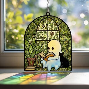May include: A stained glass suncatcher featuring a white ghost reading an open book next to a black cat. The scene is set within a green arched window frame filled with leafy vines, flowers, and a butterfly, hanging by a silver chain.