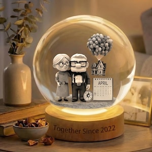 Op de afbeelding: Een sneeuwbol met een beeldje van een stel, een huis en een tros ballonnen. De houten basis heeft de tekst "Together Since 2022". Een kalender toont de maand april. De bol wordt verlicht met een warme gloed.