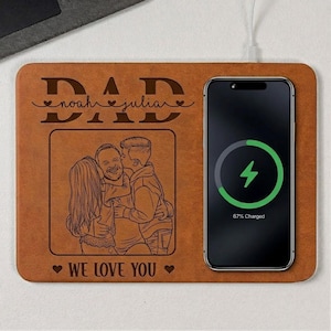 May include: A brown wireless phone charger with a personalized design. The charger features the word "DAD" and the names "noah" and "julian" engraved on the top. A photo is engraved below the names, and the words "WE LOVE YOU" are at the bottom. A phone is charging on the right.