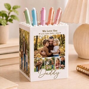 May include: A white pen holder with a photo collage and the word "Daddy". The holder contains pens in green, blue, pink, orange, and pink. The text "We Love You" and names are also visible. A personalized gift.