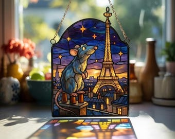 May include: Stained glass window art featuring a blue rat gazing at the Eiffel Tower in Paris. The artwork has a night sky background with stars and a gold chain for hanging. The window art is a decorative piece.