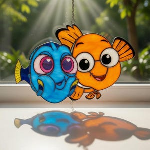 May include: A stained glass window decoration depicting two cartoon fish. One is blue with pink eyes, the other orange with large white eyes. Both fish have smiling faces and are suspended from a chain. The window decoration is casting a shadow.