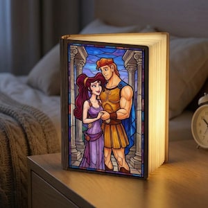 May include: A decorative book-shaped lamp featuring a stained-glass design of Hercules and Megara. The lamp is open, with the pages illuminated, and sits on a wooden nightstand. The artwork depicts the couple in a classical setting, with columns and a blue sky.