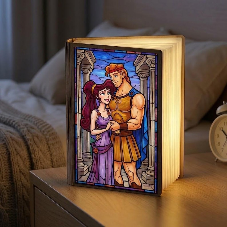 May include: A decorative book-shaped lamp featuring a stained-glass design of Hercules and Megara. The lamp is open, with the pages illuminated, and sits on a wooden nightstand. The artwork depicts the couple in a classical setting, with columns and a blue sky.