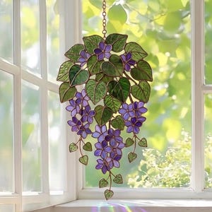 Pu&ograve; includere: Appeso per finestra in vetro colorato con un grappolo di fiori viola e foglie verdi. Il design floreale &egrave; sospeso da una catena. L'arte della finestra &egrave; esposta davanti a una finestra con luce naturale.