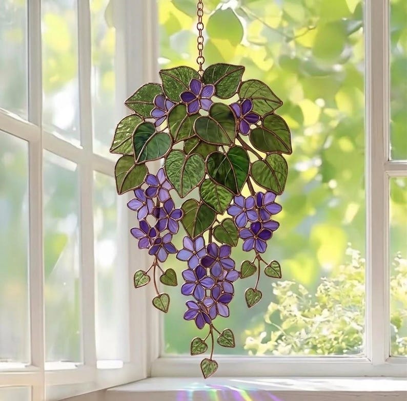 Pu&ograve; includere: Appeso per finestra in vetro colorato con un grappolo di fiori viola e foglie verdi. Il design floreale &egrave; sospeso da una catena. L'arte della finestra &egrave; esposta davanti a una finestra con luce naturale.
