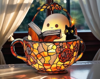 May include: A stained glass tea cup lamp featuring warm orange and yellow floral patterns. Inside the cup sits a white ghost figure reading an open book, accompanied by a small black cat with yellow eyes. The lamp has a decorative handle.