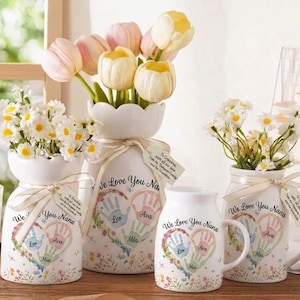 May include: A collection of white ceramic vases and mugs, each adorned with a heart-shaped handprint design and the text "We Love You Nana." The vases hold colorful flowers, and the mugs have handles. Each piece is tied with a ribbon.
