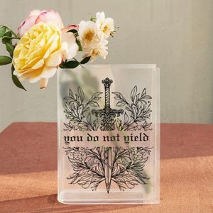 May include: Clear rectangular vase with a black sword and floral design, and the text "you do not yield". The vase is filled with yellow and white roses, set on a pink surface.