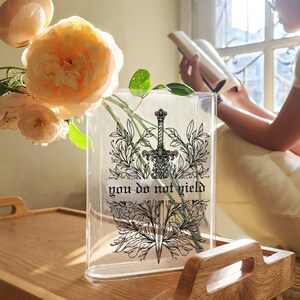 May include: A clear rectangular vase with a black and white sword design and the text "you do not yield." The vase holds flowers and is on a wooden tray. The flowers are peach-colored.