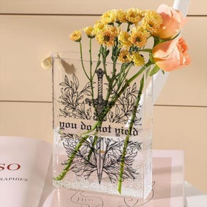 May include: A clear rectangular vase with a black sword and floral design, holding yellow chrysanthemums and peach roses. The vase has the text "you do not yield". The flowers are arranged in water.