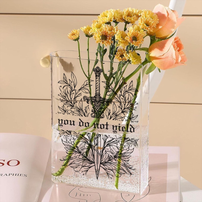 May include: A clear rectangular vase with a black sword and floral design, holding yellow chrysanthemums and peach roses. The vase has the text "you do not yield". The flowers are arranged in water.
