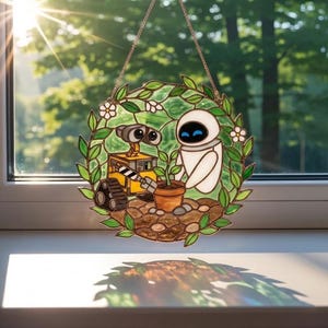 May include: Stained glass window art featuring the characters Wall-E and Eve from the animated film. The artwork depicts the robots with a plant in a pot, surrounded by green leaves and white flowers. The piece is round and hangs from a gold chain.