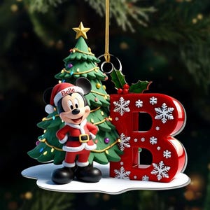 Può includere: Un addobbo natalizio con un topo dei cartoni animati che indossa un costume e un cappello da Babbo Natale con il nome Belle stampato, accanto a un albero di Natale verde decorato e una grande lettera B rossa con motivi a fiocchi di neve.