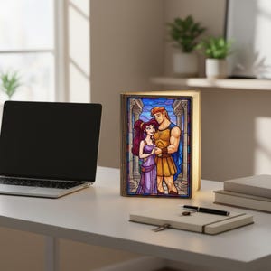 May include: A decorative book-shaped lamp featuring a stained-glass style illustration of Hercules and Megara. The lamp has a warm, inviting glow and is set on a white desk. The artwork depicts the couple in a romantic embrace, with a classical architectural background.
