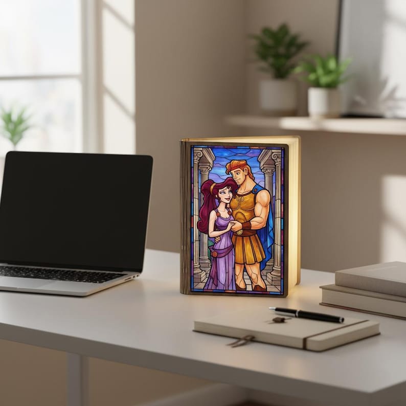 May include: A decorative book-shaped lamp featuring a stained-glass style illustration of Hercules and Megara. The lamp has a warm, inviting glow and is set on a white desk. The artwork depicts the couple in a romantic embrace, with a classical architectural background.