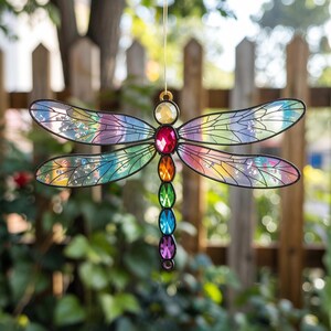 May include: A colorful dragonfly suncatcher with iridescent wings and a black outline. The dragonfly features a series of jewel-toned gems in red, orange, green, blue, and purple. The wings are a mix of rainbow colors.