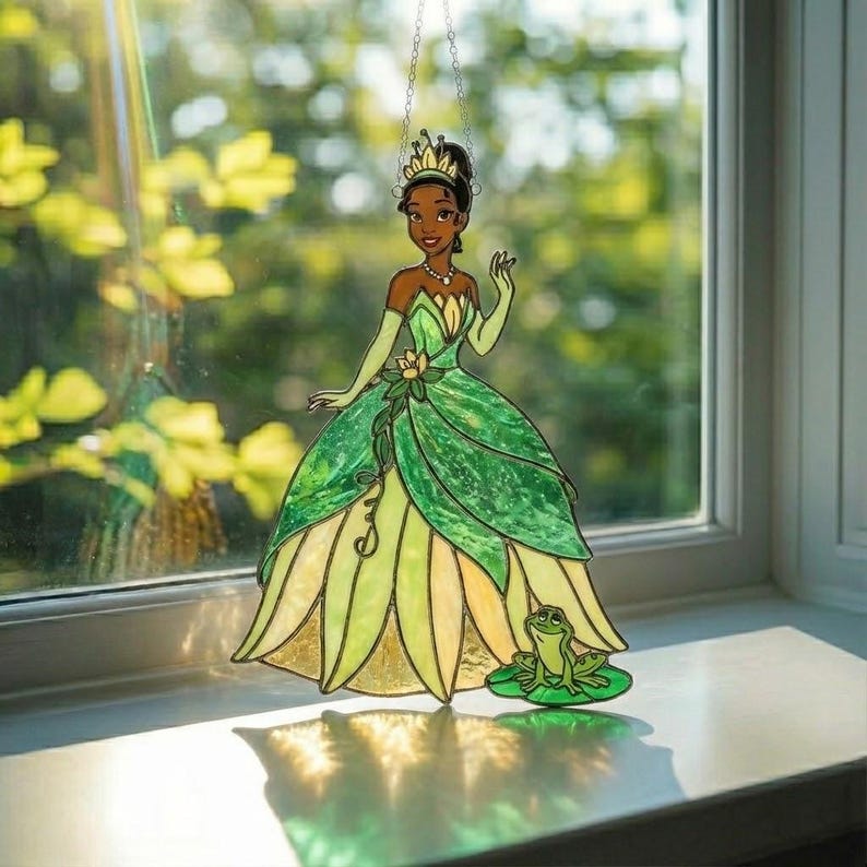 K&ouml;nnte beinhalten: Ein Buntglas-Suncatcher, der eine dunkelh&auml;utige Figur in einem gr&uuml;nen Kleid mit goldener Krone darstellt, erg&auml;nzt durch einen kleinen gr&uuml;nen Frosch auf einem Seerosenblatt am Saum des Kleides, aufgeh&auml;ngt an einer d&uuml;nnen Metallkette.