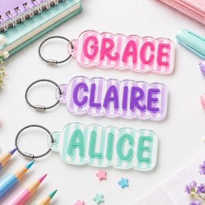 Puede incluir: Tres llaveros rectangulares de acrílico con bordes festoneados y rayas verticales. El llavero superior es rosa con el nombre GRACE en rosa oscuro, el del medio es morado con CLAIRE en morado oscuro y el inferior es verde azulado con ALICE en verde.