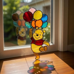 Könnte beinhalten: Buntglasfenster von Winnie Puuh, der bunte Luftballons hält. Der gelbe Bär trägt ein rotes Hemd. Die Ballons sind rot, orange, gelb, blau und grün. Das Bild ist vor einem Fenster mit einer Holzfensterbank platziert.