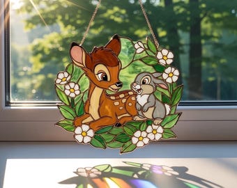 May include: Stained glass window art featuring Bambi and Thumper surrounded by white flowers and green leaves. The artwork is framed with a gold-colored metal and hangs from a delicate chain. The window art is displayed in front of a bright, sunny window.