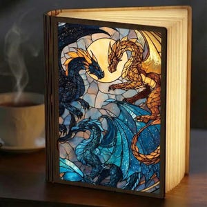 May include: A wooden book-shaped lamp with a stained-glass design featuring three dragons in shades of blue, gold, and black against a moonlit background. The lamp is open, with the pages illuminated, and sits on a dark wooden surface.