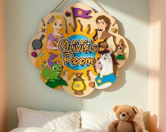 Puede incluir: Un letrero de madera con forma de flor con el texto "Olivia's Room". El letrero presenta personajes de dibujos animados y un diseño de sol. Cuelga de una pared sobre una cama con un oso de peluche.