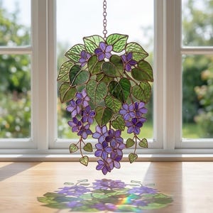 Pu&ograve; includere: Un elemento decorativo a sospensione in vetro colorato con fiori viola e foglie verdi. Il design floreale &egrave; sospeso da una catena davanti a una finestra. La luce solare proietta un'ombra colorata sulla superficie in legno.