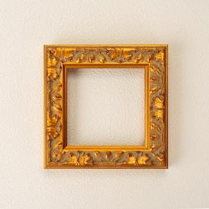 Puede incluir: Un marco de fotos cuadrado y dorado con intrincadas tallas en relieve. El marco tiene un acabado dorado antiguo y presenta un patrón repetido de hojas y otros elementos decorativos. El marco está vacío, listo para exhibir una foto o una obra de arte.