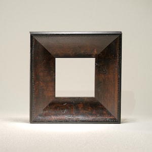 May include: A square, dark brown wooden picture frame with a lighter square opening in the center. The frame has a decorative edge and a slightly angled design, creating a shadow effect. The wood grain is visible.