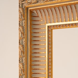 May include: Close-up of a decorative picture frame with a gold and off-white color scheme. The frame features ornate detailing, including a gold inner border and a textured, ribbed design on the off-white sections. The frame's corner is visible.