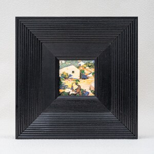 May include: A square painting featuring a landscape with a small white house, set within a wide, black, ribbed wooden frame. The artwork displays a sunlit scene with earthy tones, green foliage, and blue shadows, encased in a deep, textured border.