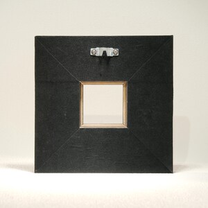 May include: Back of a square black framed artwork with a metal hanger attached. The frame has a square opening with a light-colored wood frame. The black frame has a textured appearance.