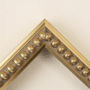 May include: A corner section of a picture frame featuring a metallic gold finish. The frame has a beveled outer edge and an inner border decorated with a repeating pattern of raised, rounded bead-like spheres that follow the mitered corner joint.